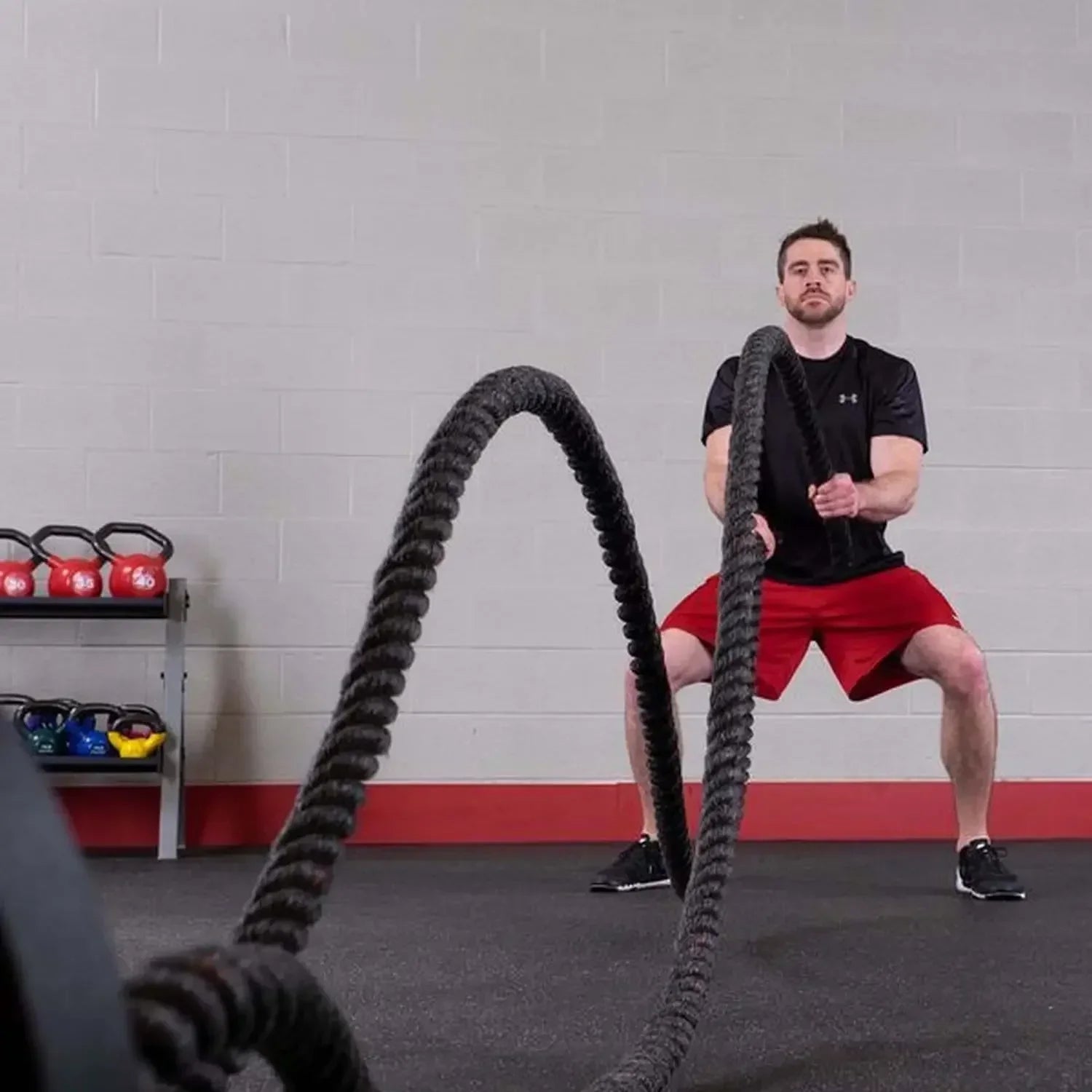 Mann trainiert mit Battle Ropes im Fitnessstudio, Krafttraining mit Kettlebells im Hintergrund