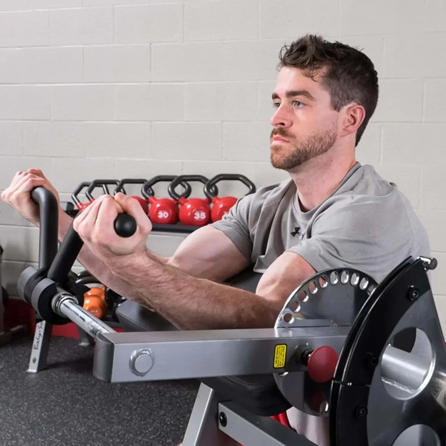 Mann trainiert mit Kraftstation im Fitnessstudio, rote Kettlebells im Hintergrund